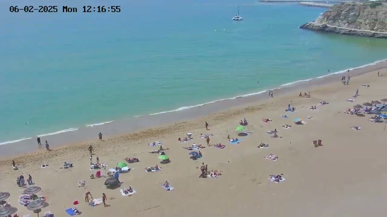 A sandy beach with many people sunbathing and swimming in calm, turquoise water under a clear sky.
