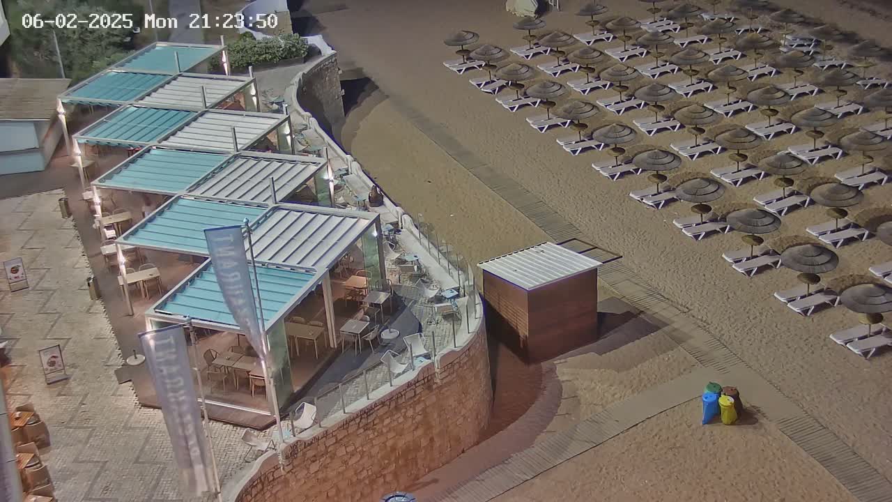An outdoor nighttime scene shows a restaurant with multiple covered patio areas next to a sandy beach with rows of lounge chairs and umbrellas.