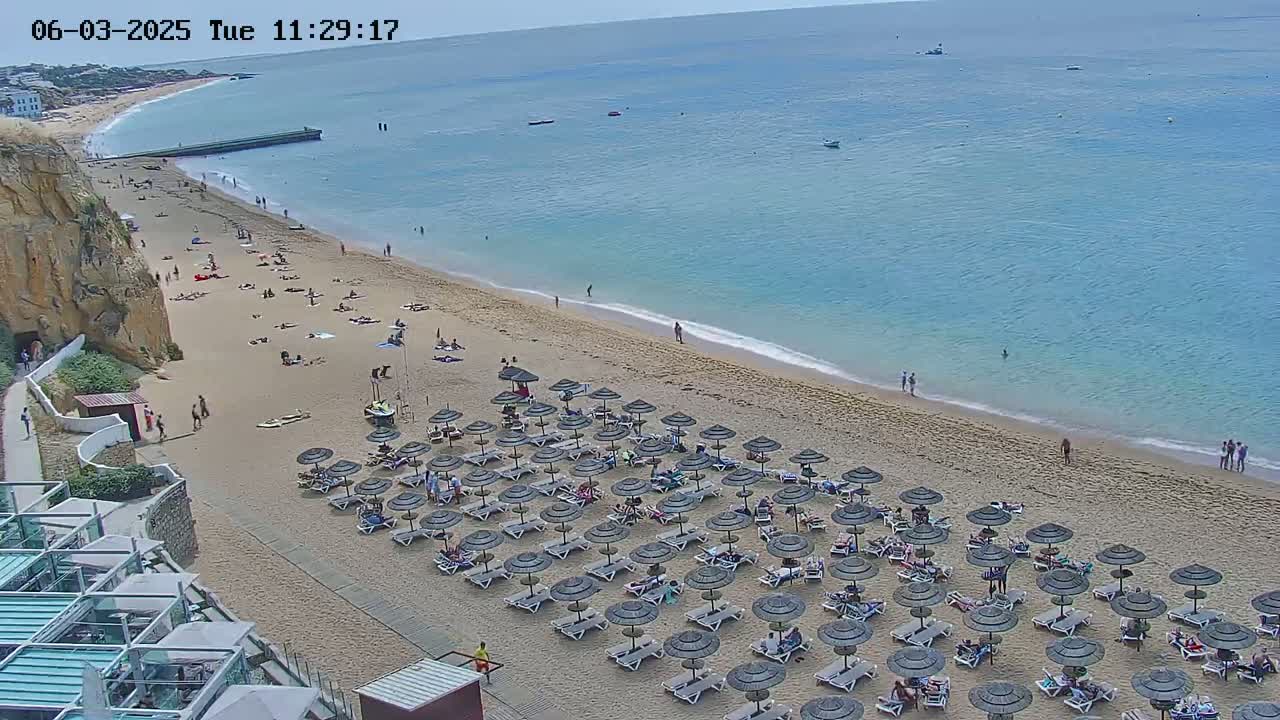A sandy beach with numerous umbrellas and lounge chairs is partially populated by people on a partly sunny day, next to a calm ocean.