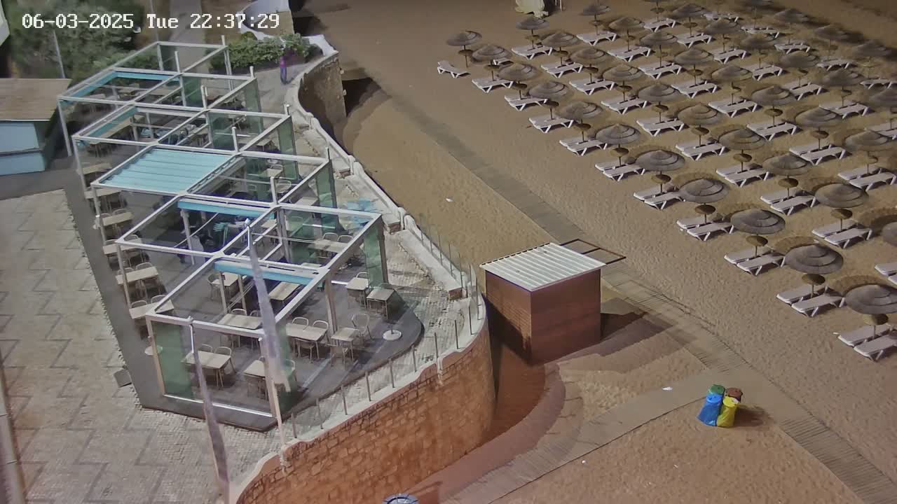 An outdoor, nighttime scene shows a beach with many lounge chairs and umbrellas next to a restaurant with glass-walled patio seating.