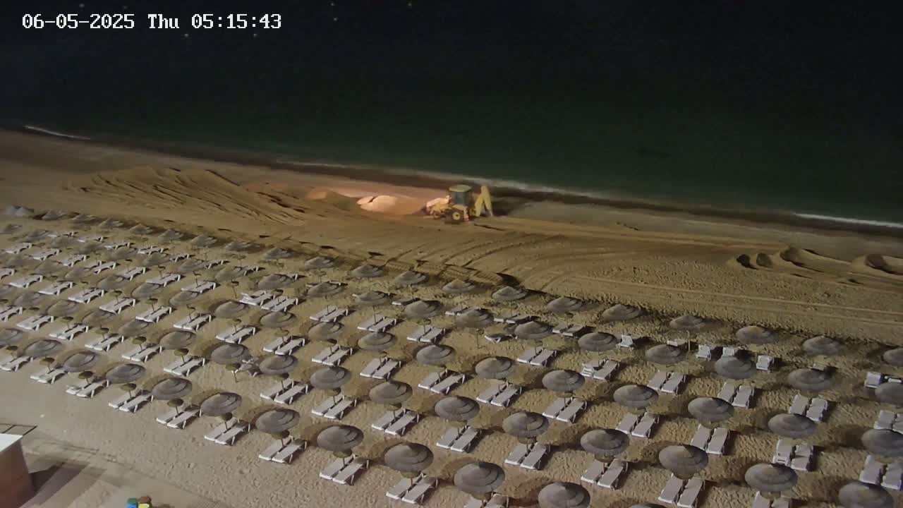 At night, a beach with rows of closed umbrellas and lounge chairs is being worked on by a piece of heavy machinery near the water's edge.