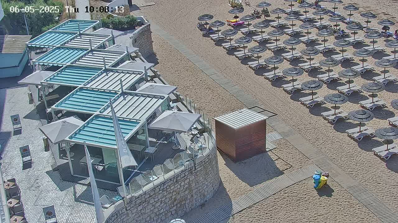 An outdoor scene on a sunny day shows a row of beach umbrellas and lounge chairs next to a stone wall with a series of covered outdoor seating areas.