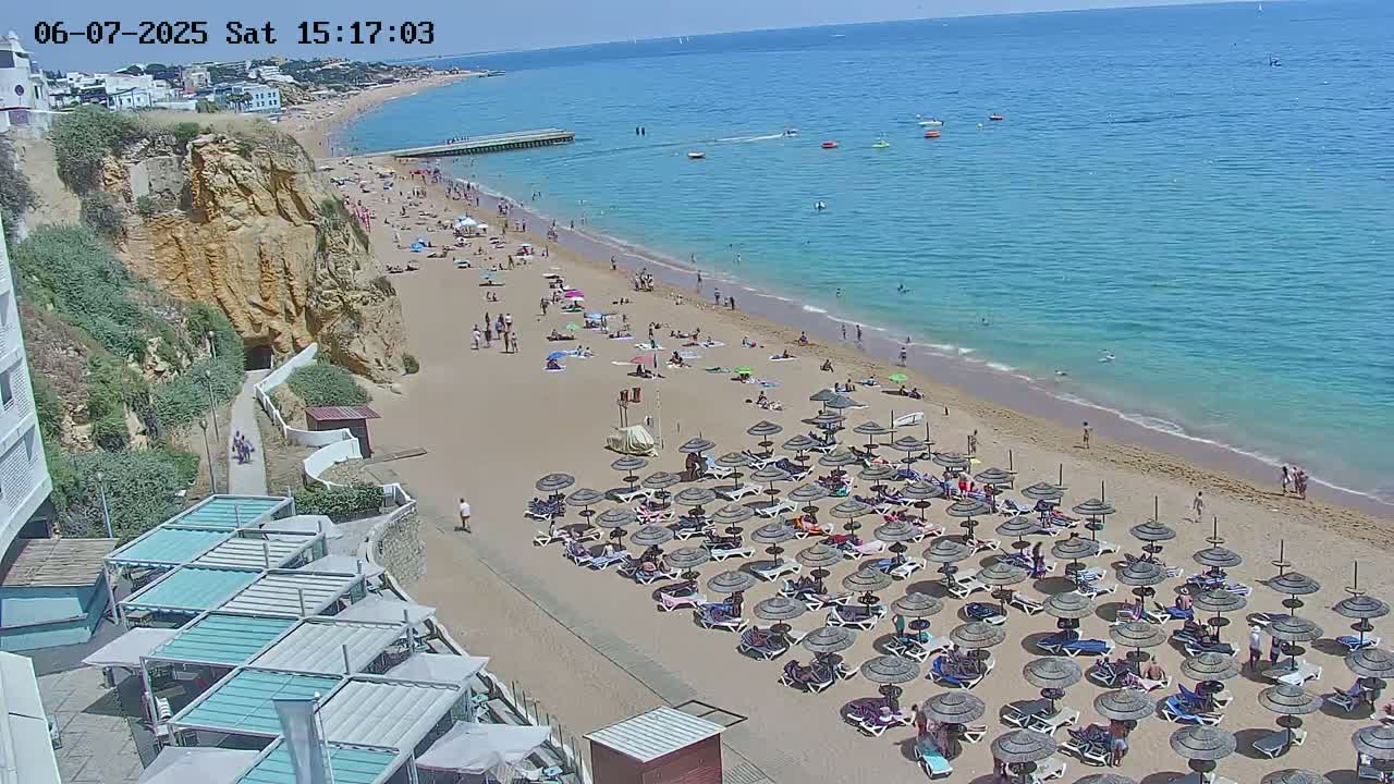 A sandy beach with numerous umbrellas and beachgoers stretches along a calm, clear blue ocean under a sunny sky, backed by a high cliff and buildings.