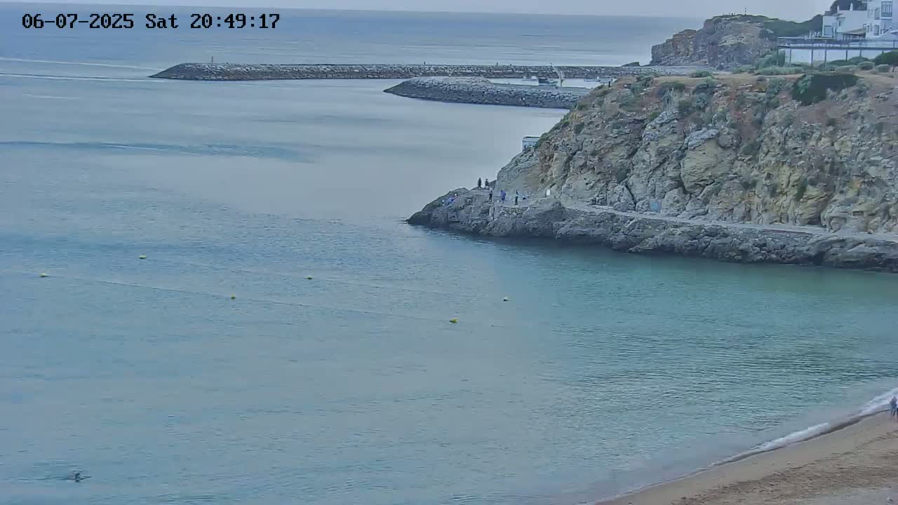 A calm, light-blue ocean bay is partially enclosed by a rocky cliff, with a sandy beach at the bottom right, and several people are visible near the cliff's edge.