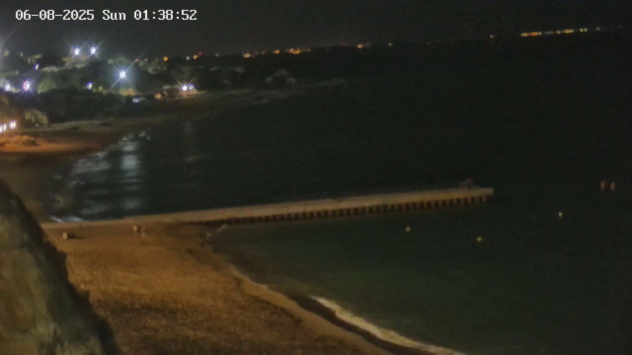 A nighttime view of a sandy beach with a pier extending into dark water, under a clear sky with lights visible in the distance.