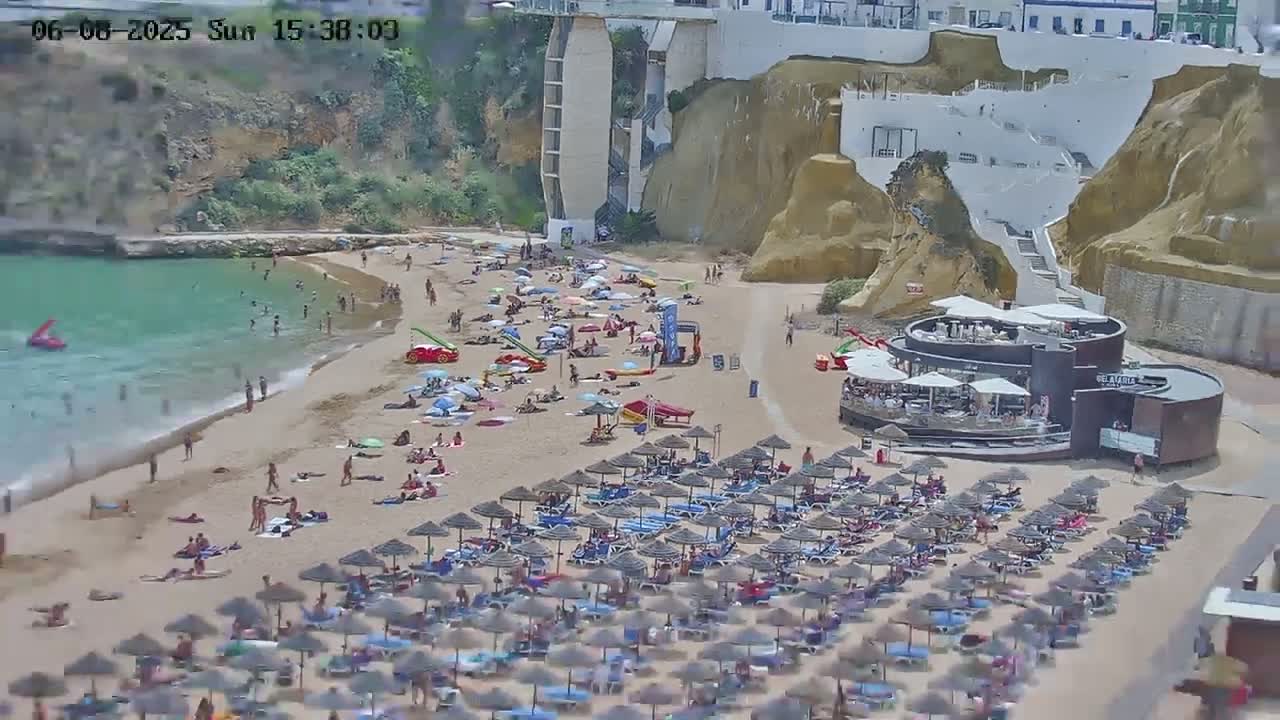 A sandy beach filled with people under numerous umbrellas, near a building and cliffs, on a sunny day.