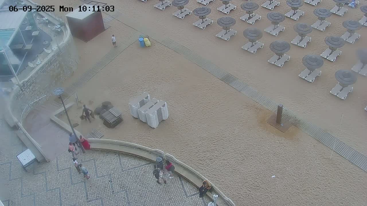 A sandy beach with numerous closed umbrellas and lounge chairs is shown, with several people near a building and walkway; the weather appears overcast.