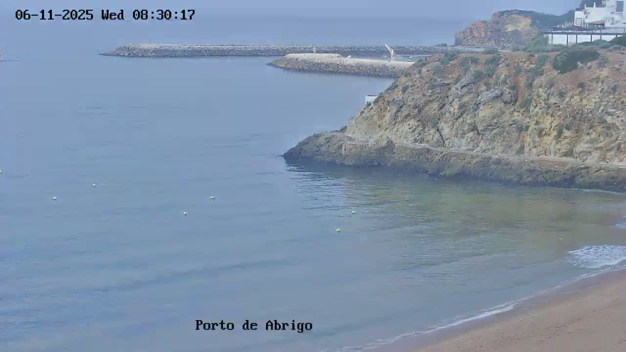A calm, hazy ocean bay is seen from a high vantage point, showing a sandy beach, rocky cliffs, a breakwater, and several small buoys in the water.