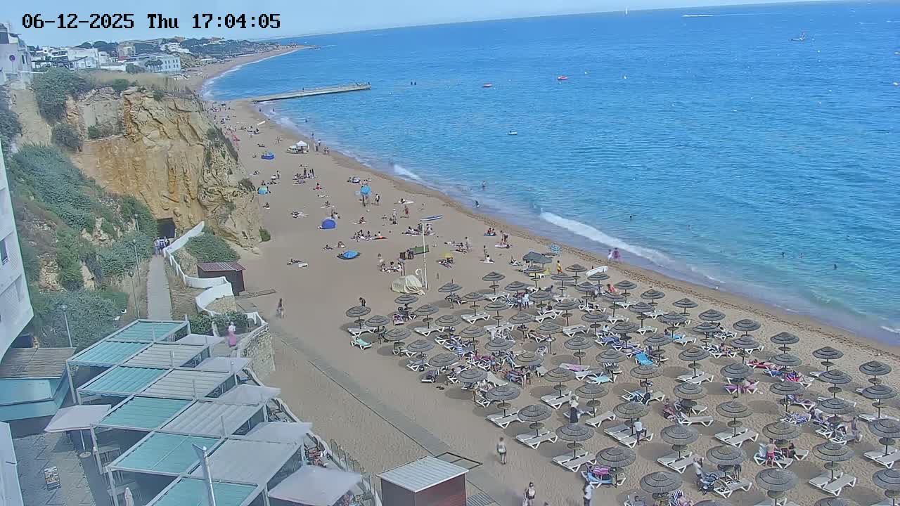 A sandy beach with numerous dark umbrellas and sunbathers stretches along a clear blue ocean under a sunny sky, adjacent to a high cliff face and some buildings.
