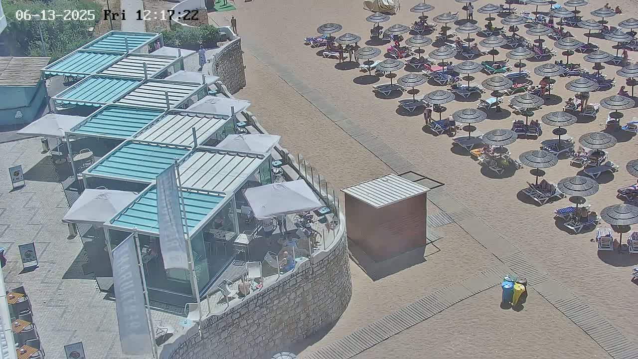 An aerial view shows a sandy beach with numerous dark umbrellas and lounge chairs, adjacent to a building with multiple teal-roofed outdoor structures.