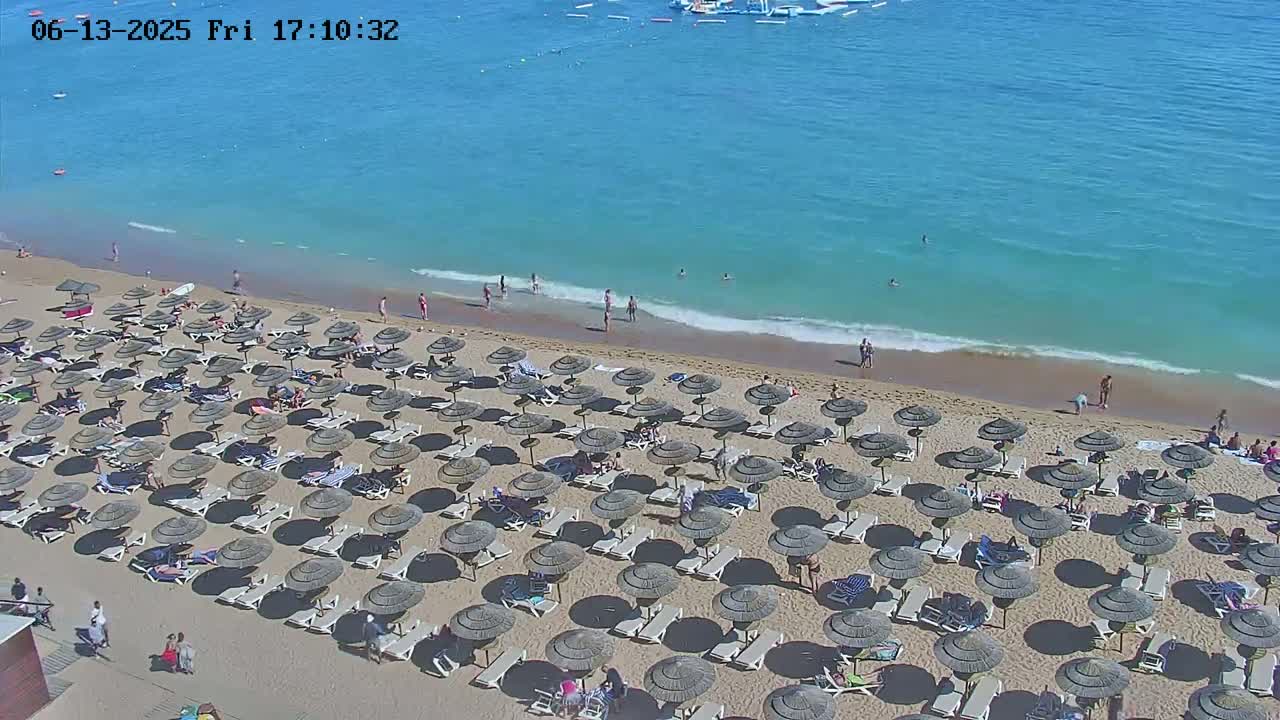 A sandy beach with numerous thatched umbrellas and lounge chairs is populated by bathers and sunbathers, under a sunny sky, next to a calm, clear turquoise ocean.