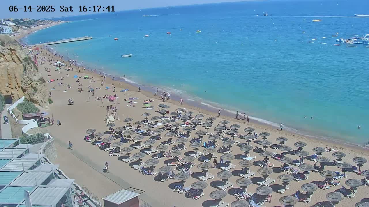 A sandy beach with numerous umbrellas and lounge chairs is crowded with people, adjacent to a clear blue ocean on a sunny day.