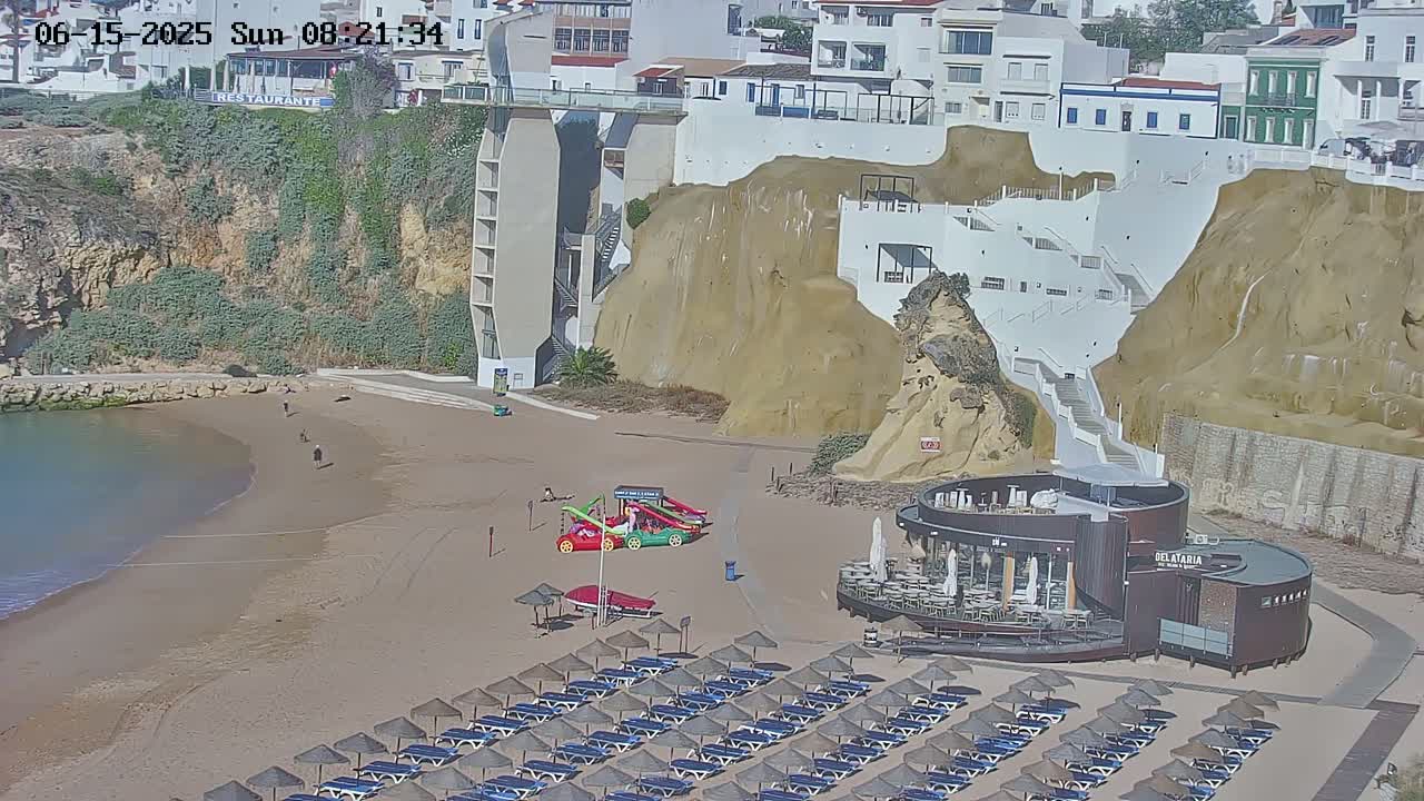 A sandy beach with rows of beach umbrellas and chairs, a small restaurant, and children's play equipment sits below a cliffside with buildings and stairs on a sunny day.