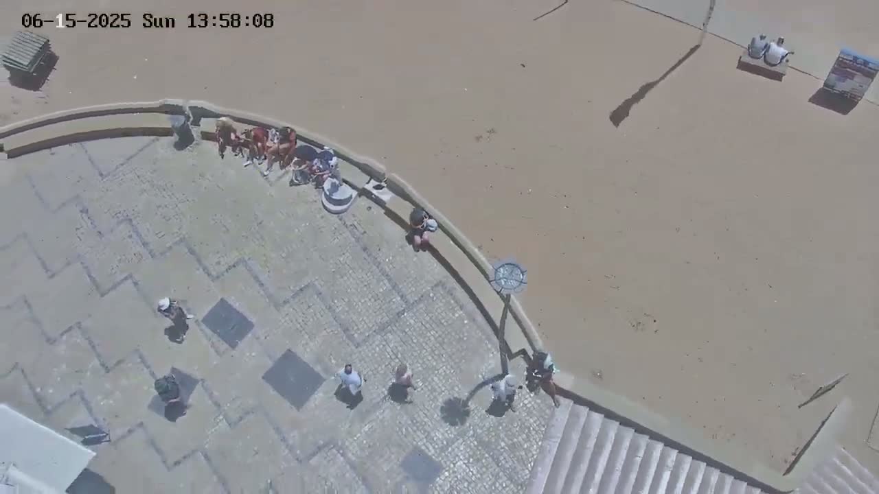 A paved area with several people sitting and walking near a sandy beach on a sunny day.