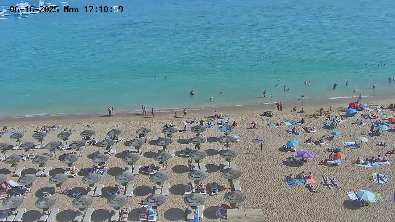 A sandy beach with numerous umbrellas and sunbathers is adjacent to a calm, turquoise ocean where people are swimming.