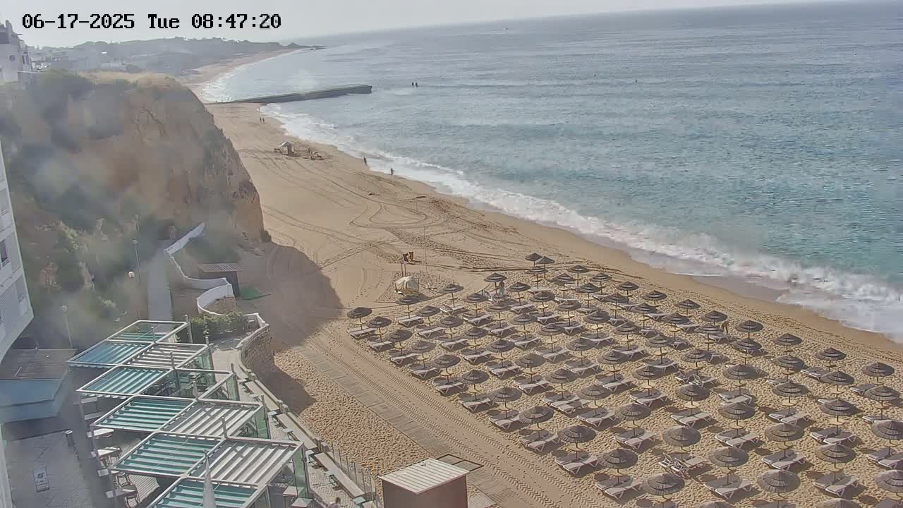 A sandy beach with numerous umbrellas and lounge chairs sits below a cliffside on a partly cloudy day, next to a calm ocean with gentle waves.