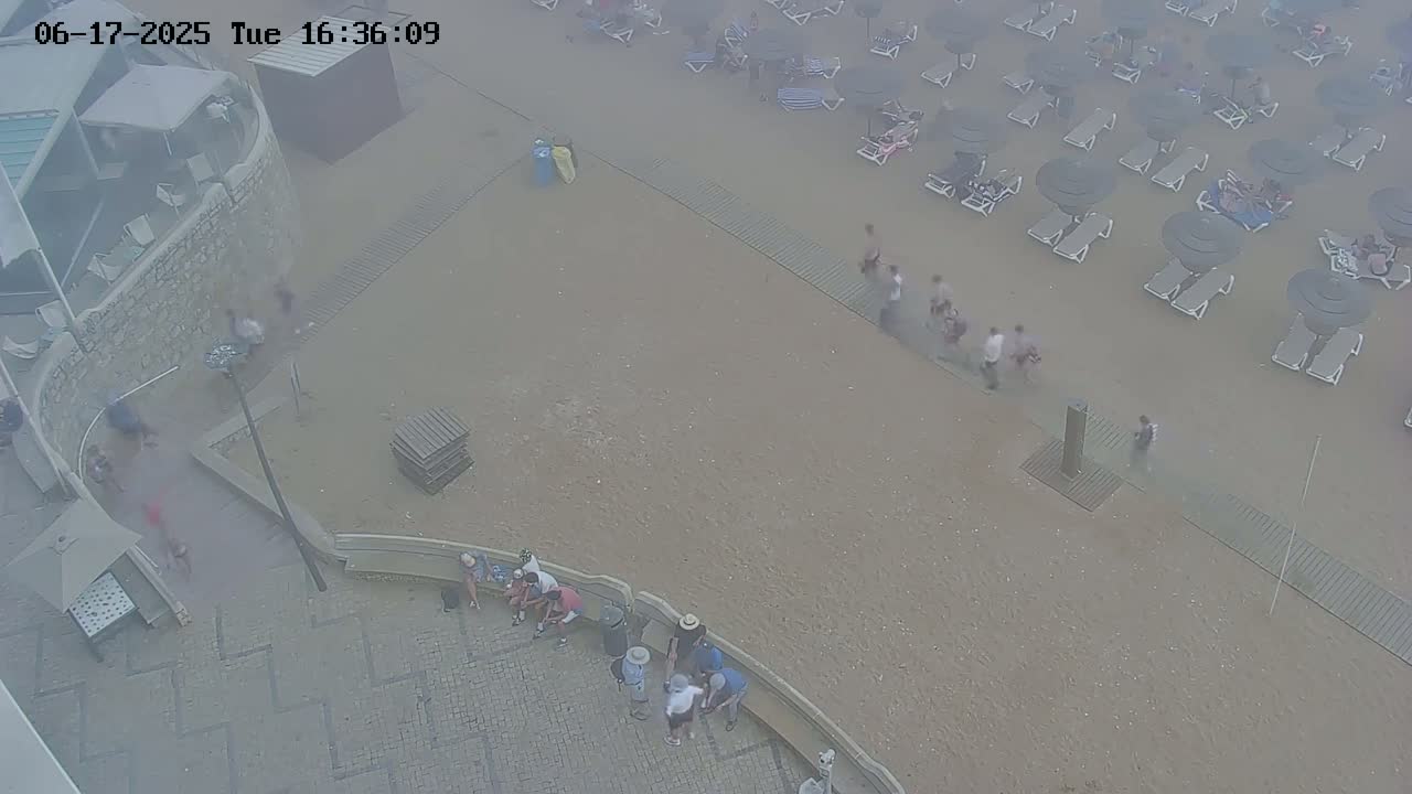 A hazy, high-angle view shows a beach with people scattered about, some under umbrellas and lounge chairs, and others near a stone wall and paved area.