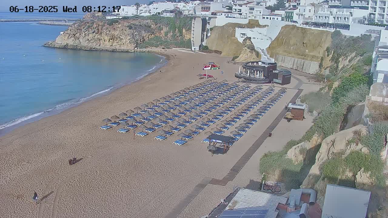 A sandy beach with numerous rows of closed umbrellas and lounge chairs sits below a cliffside of white buildings, overlooking a calm, light-blue ocean on a sunny day.