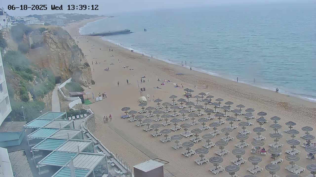 A sandy beach with numerous dark umbrellas and lounge chairs stretches along a calm, hazy ocean under an overcast sky, adjacent to a cliffside with buildings.