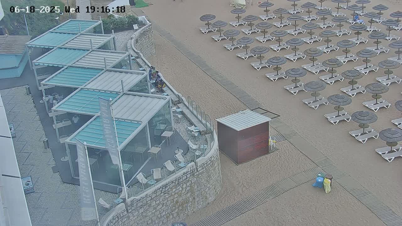 An outdoor restaurant with glass walls and a patio overlooks a sandy beach with numerous closed umbrellas and lounge chairs, under overcast skies.