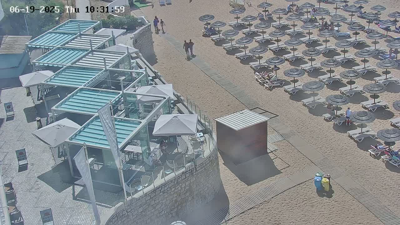 An outdoor scene shows a sandy beach with numerous umbrellas and lounge chairs, a stone wall with a patio area containing multiple covered seating areas, and several people walking.