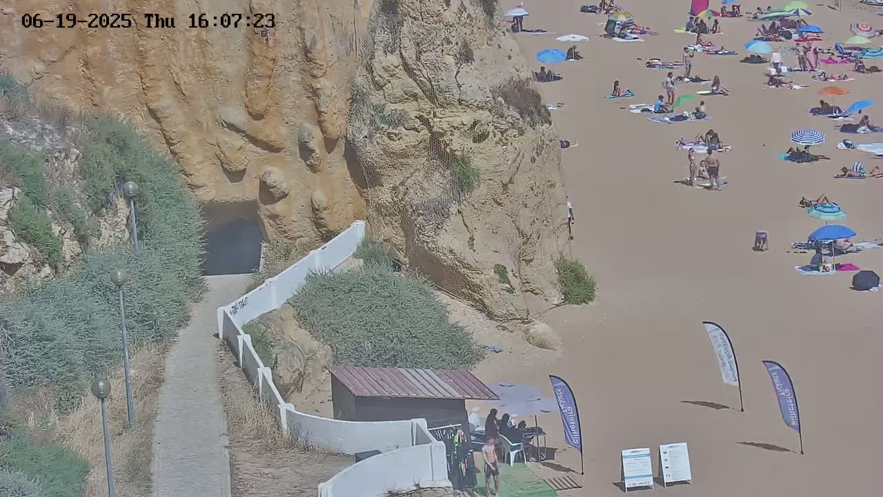 A sandy beach with many people sunbathing under umbrellas, near a path leading to a tunnel in a tan cliff face.