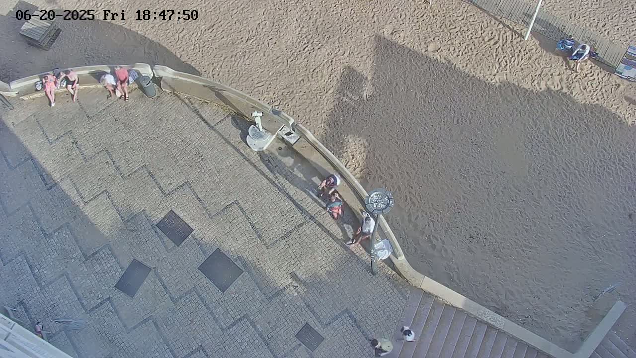 A paved area with several people sitting or standing, adjacent to a sandy beach on a sunny day.