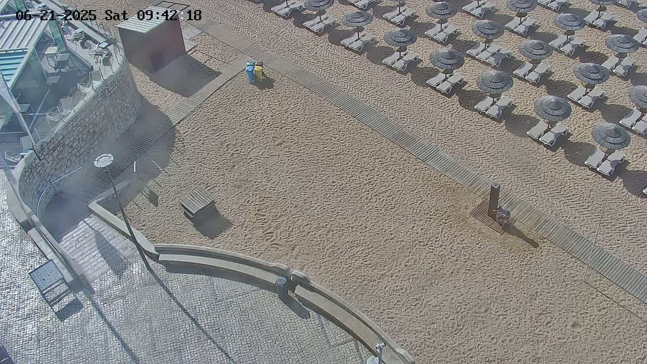 An aerial view shows a sandy beach with numerous lounge chairs and umbrellas under a sunny sky, adjacent to a paved area with stone walls and steps leading down to the beach.