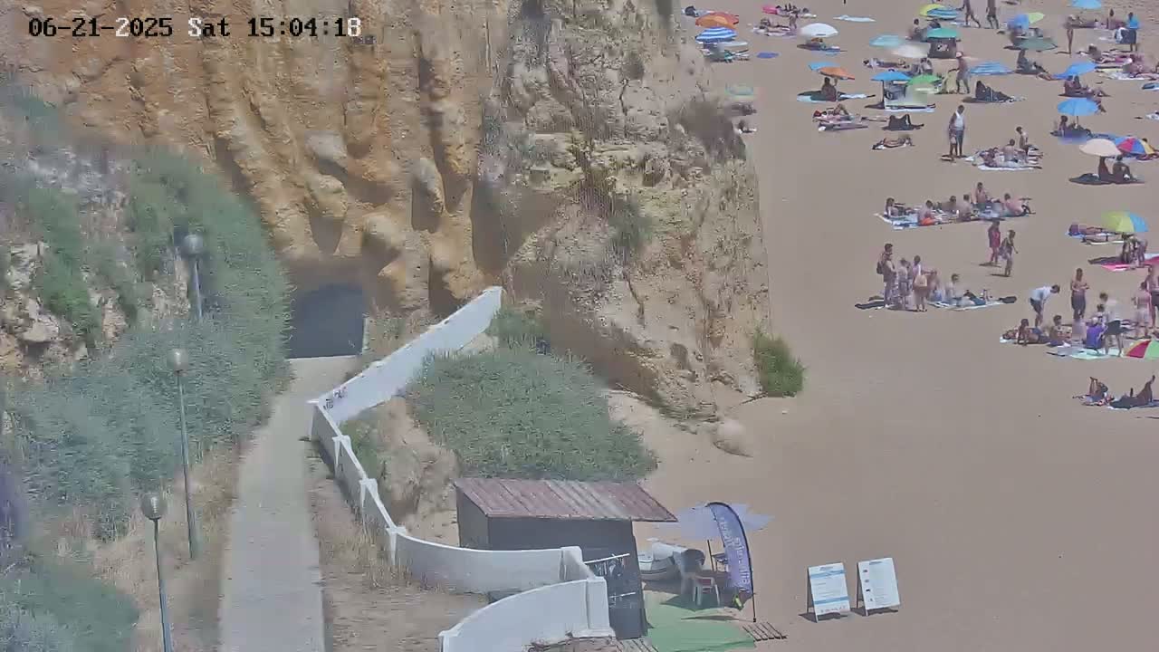 A sandy beach with numerous people sunbathing under colorful umbrellas near a cliff with a tunnel entrance, on a sunny day.