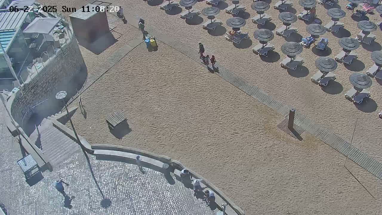 An aerial view shows a sandy beach with numerous umbrellas and lounge chairs, wooden walkways, and a few people walking near a stone staircase leading down to the beach on a sunny day.