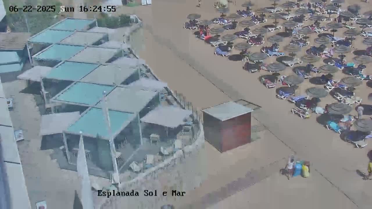 An aerial view shows a beach with numerous umbrellas and lounge chairs, adjacent to a building with multiple covered outdoor seating areas on a sunny day.