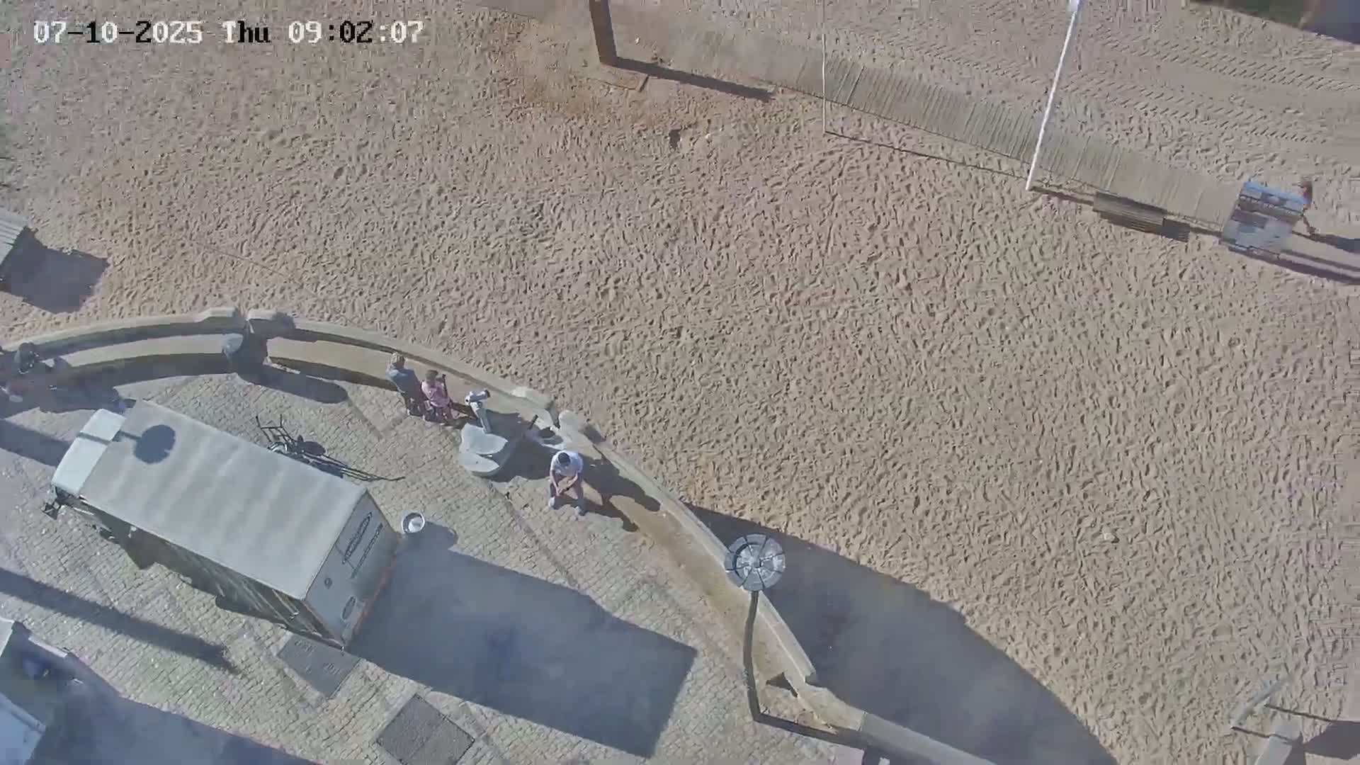 An aerial view shows a sandy area with several people sitting near a van parked on a paved surface next to a curved stone wall.