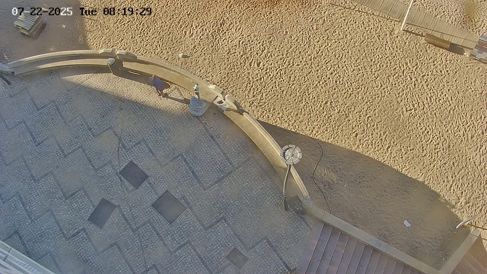An overhead view shows a sandy area next to a paved walkway with a geometric pattern, a person walking, and a small fountain.