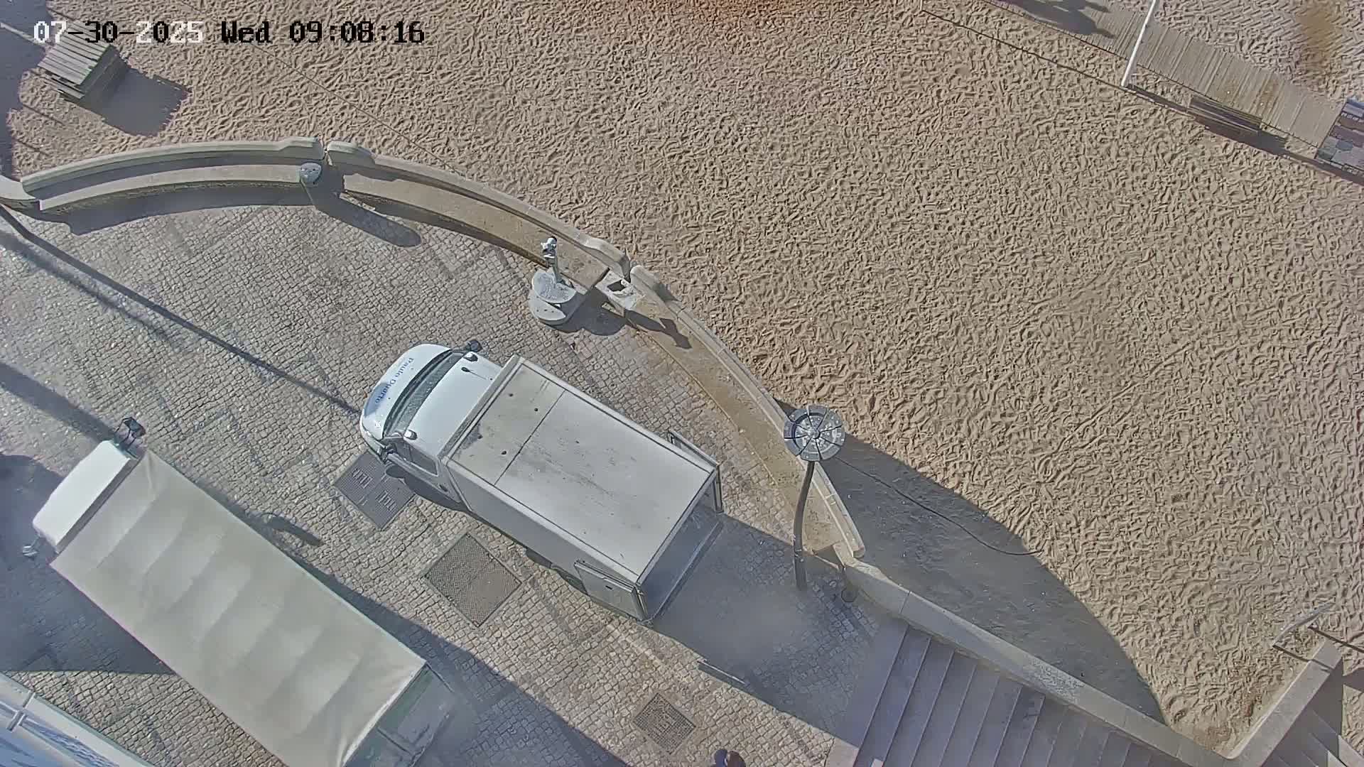 An aerial view shows a sandy beach area next to a cobblestone street where two white delivery trucks are parked, under sunny conditions.