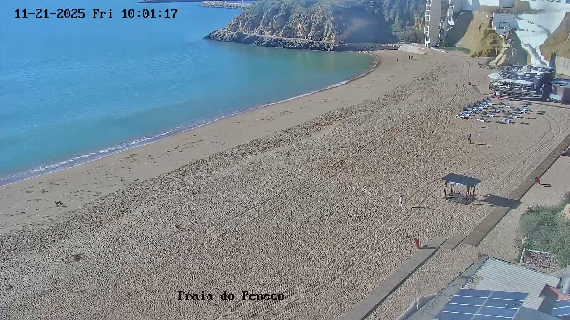 The image depicts a sunny day on a sandy beach with calm blue waters, a few people, and beachfront structures nestled against cliffs.