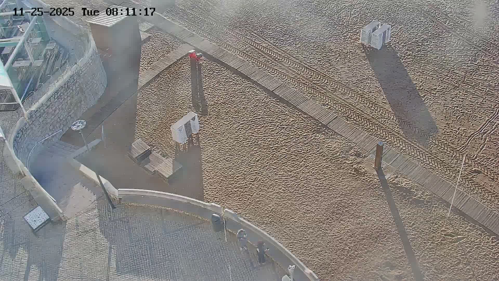 An elevated view captures a sandy beach with a wooden boardwalk and stacked chairs, bordered by a paved promenade where a few people walk, all under bright, clear conditions.