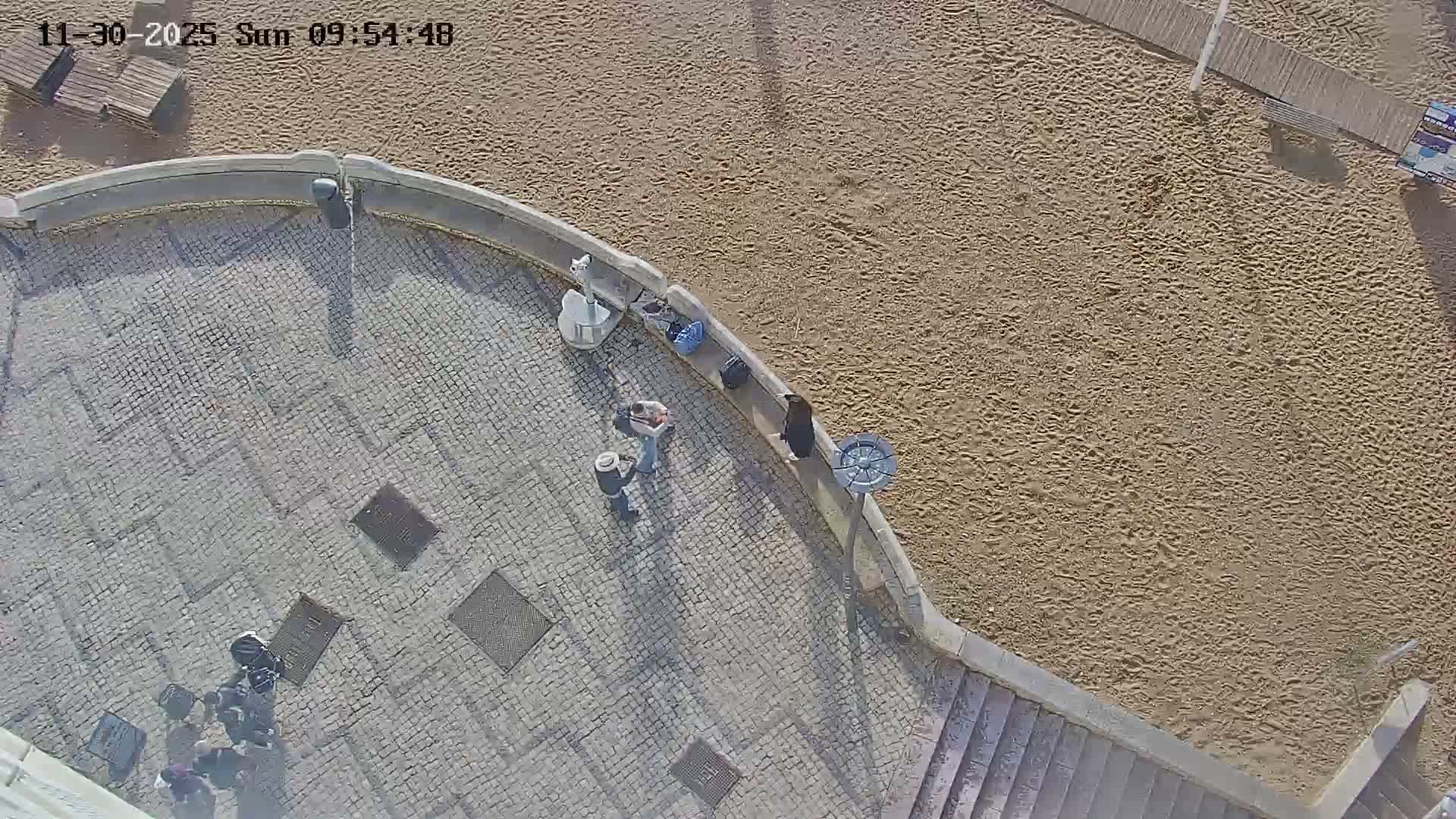 An aerial view shows a few people on a sunlit, paved promenade overlooking a sandy beach with a wooden walkway, under clear skies.