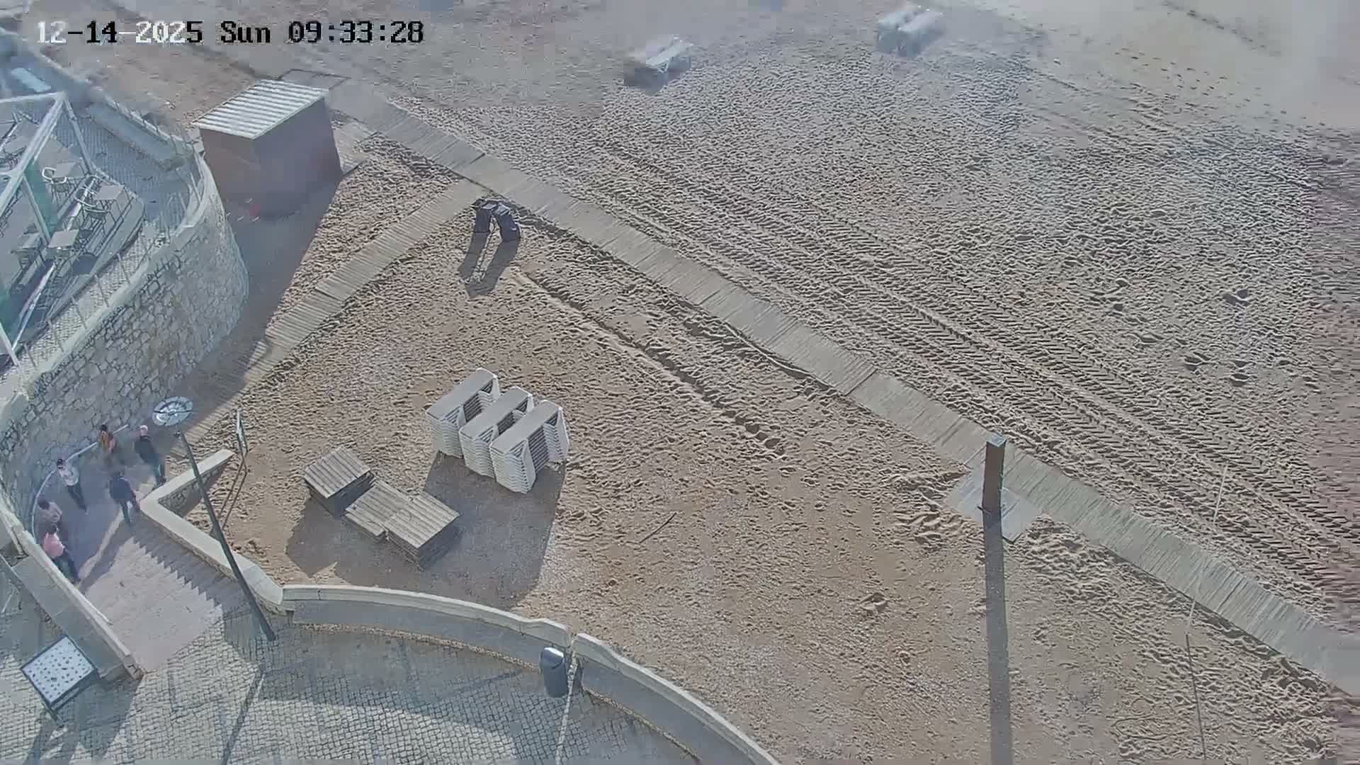 An aerial view shows a few people on a sunlit, paved promenade overlooking a sandy beach with a wooden walkway, under clear skies.
