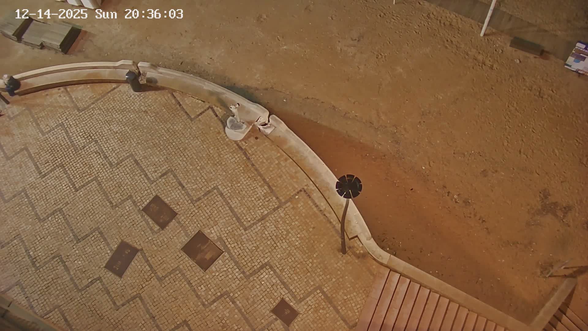 An aerial view shows a few people on a sunlit, paved promenade overlooking a sandy beach with a wooden walkway, under clear skies.