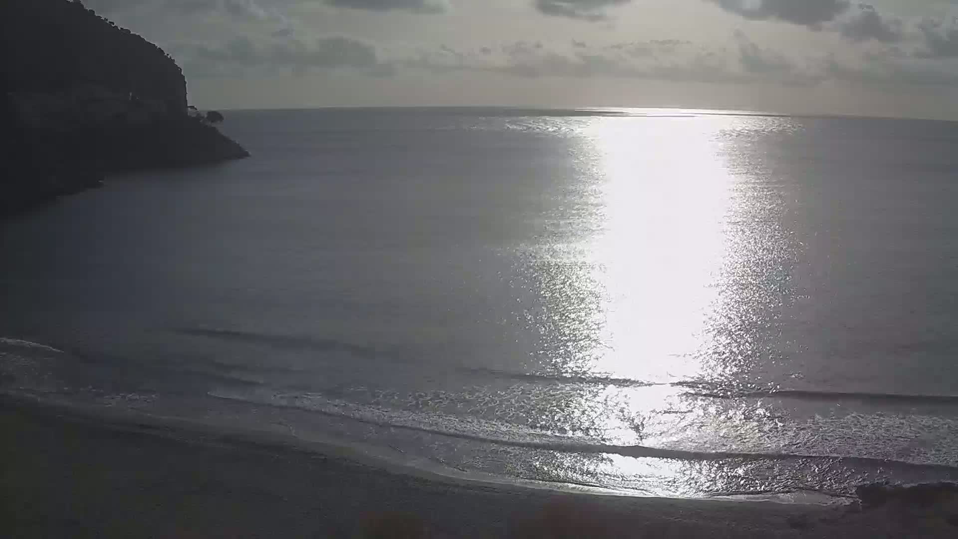 A beach with calm waves and a rocky cliff in the background under a partly cloudy sky.