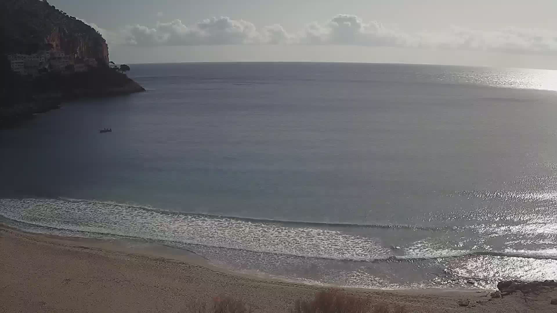 A sandy beach meets calm, gently lapping waves on a partly cloudy day, with a small boat visible in the bay and a cliff in the background.