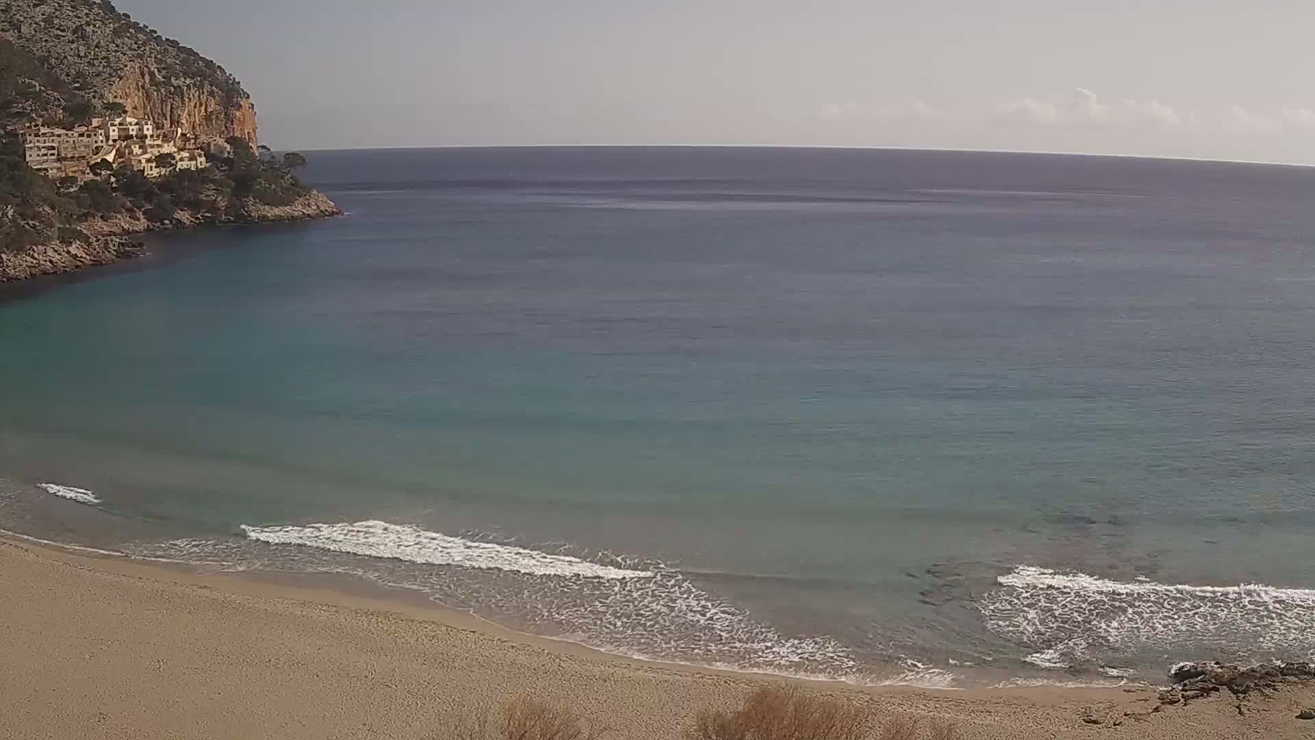 A sandy beach with small waves laps the shore of a calm, teal-colored bay nestled against a rocky cliff on a partly sunny day.