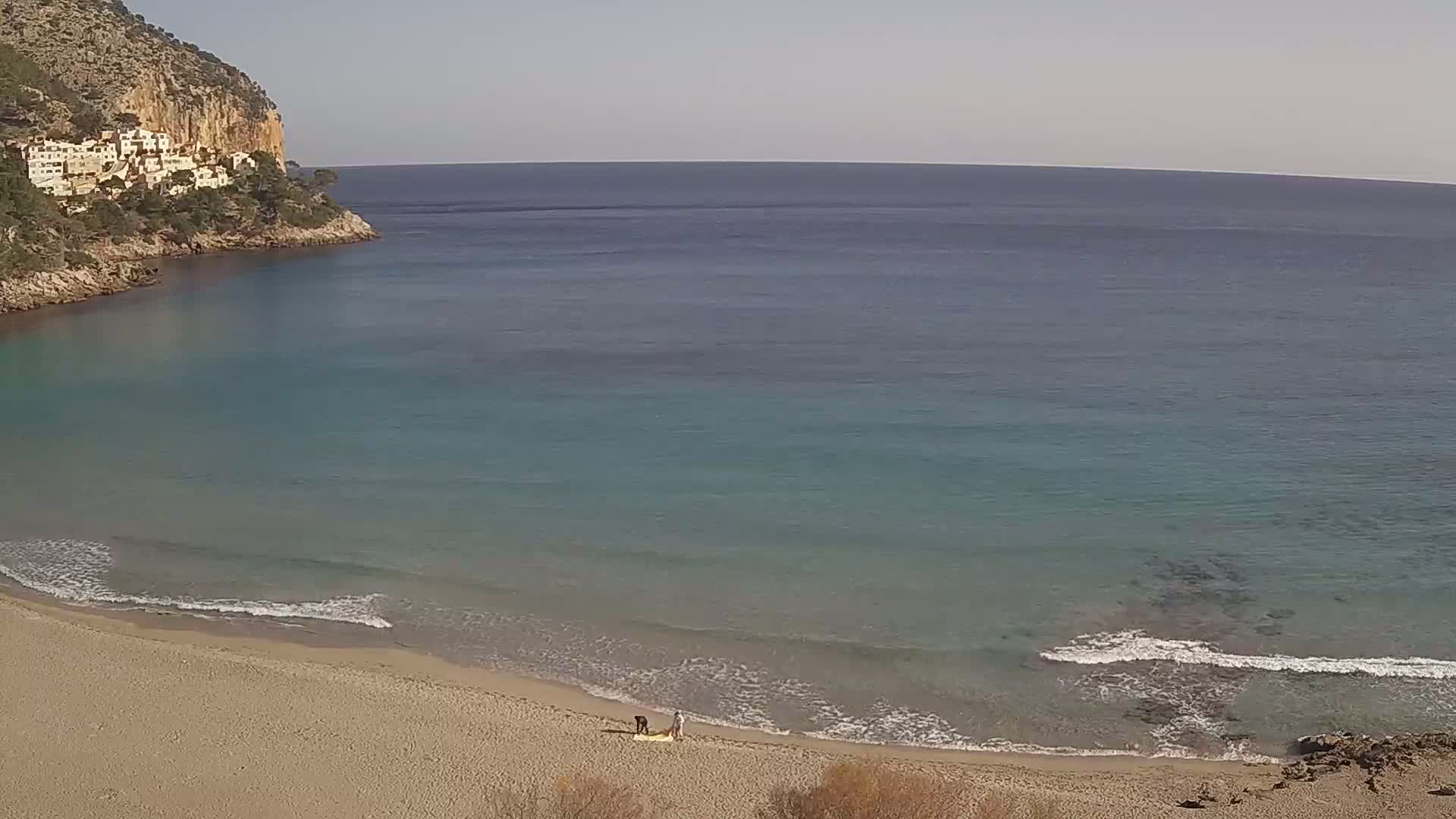 A sandy beach with calm, clear turquoise water laps gently at the shore, backed by a rocky cliff with buildings perched on its slopes under a clear sky.