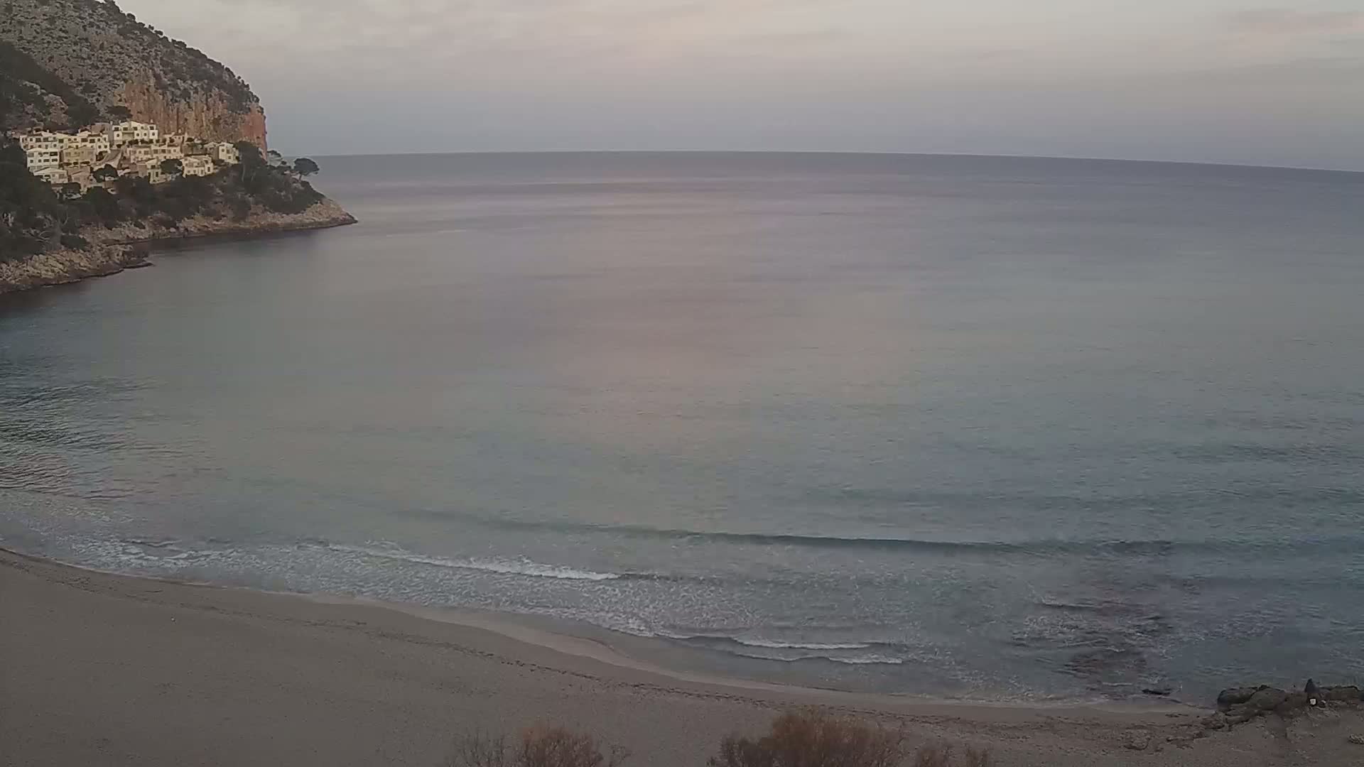 A sandy beach with gentle waves meets a calm sea under a cloudy sky, backed by a rocky cliff with buildings on top.