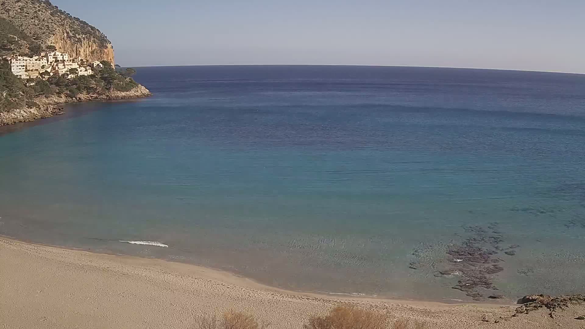 A sandy beach curves into a calm, clear turquoise sea beside a rocky cliff with buildings on top, under a bright sunny sky.