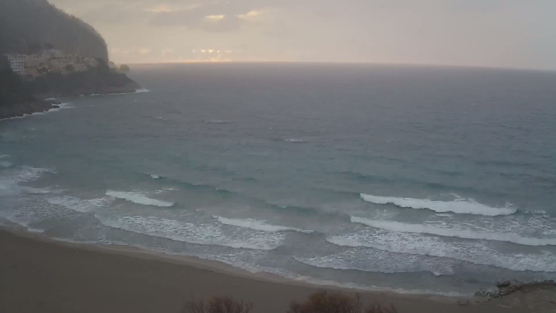 A sandy beach with small waves rolls onto shore under an overcast sky, near a rocky cliff with buildings.