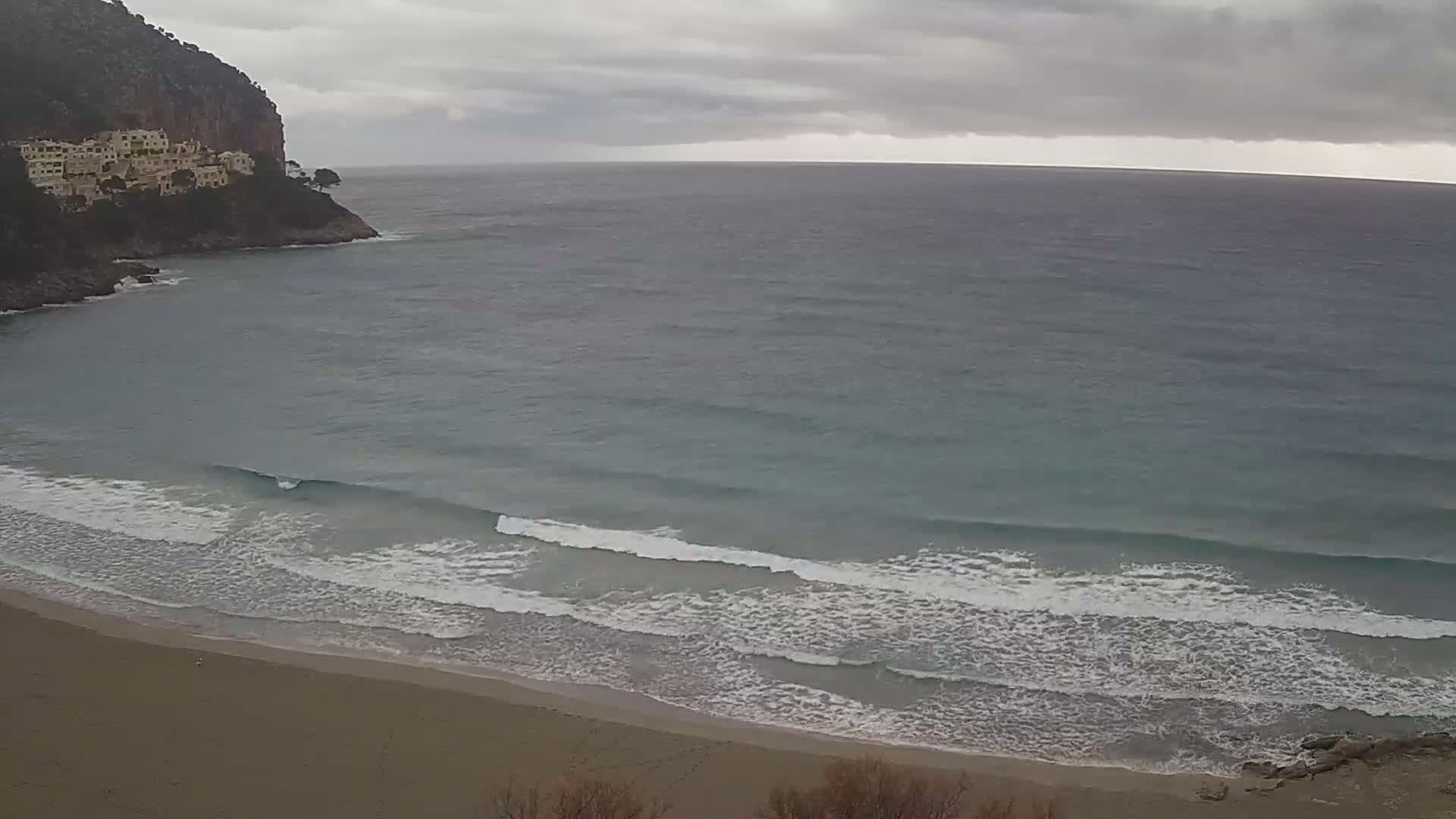 A sandy beach with small waves rolls onto the shore under an overcast sky, next to a rocky cliff with buildings.