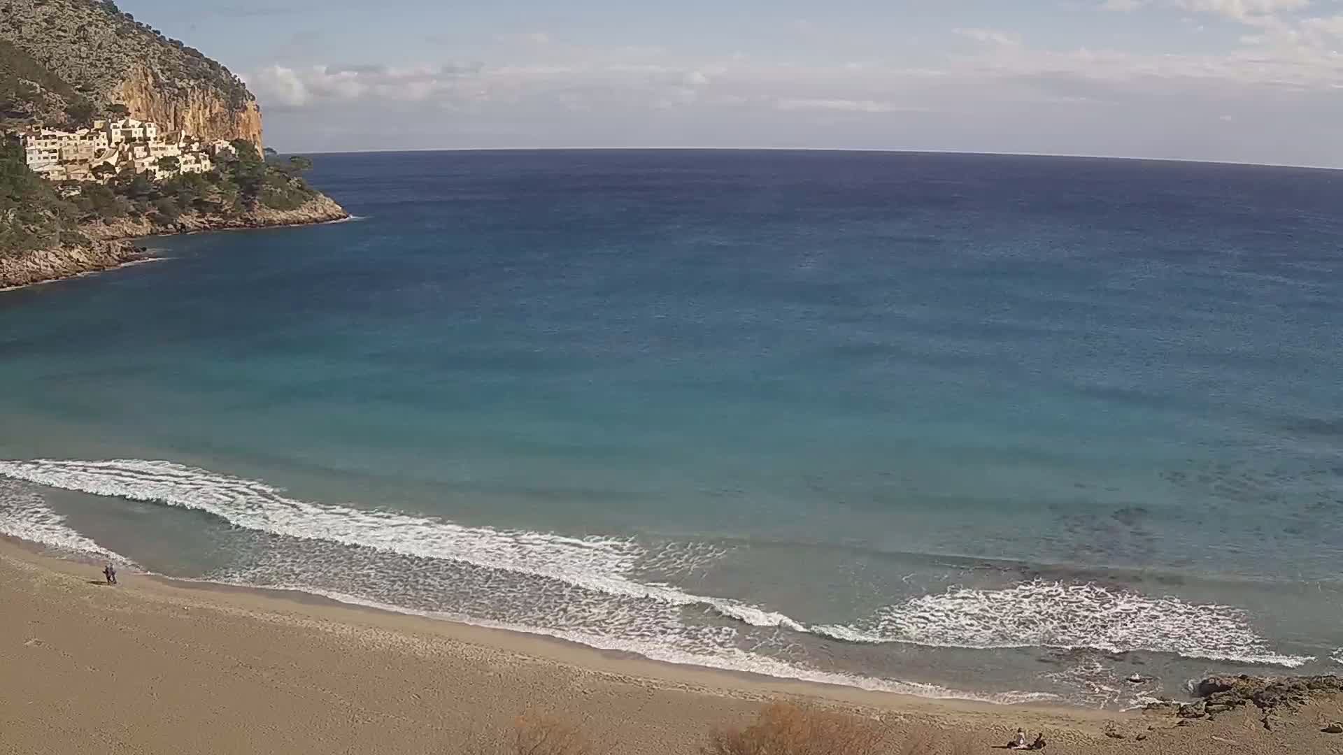 A sandy beach with gentle waves laps the shore under a partly cloudy sky, backed by a rocky cliff with buildings on top.