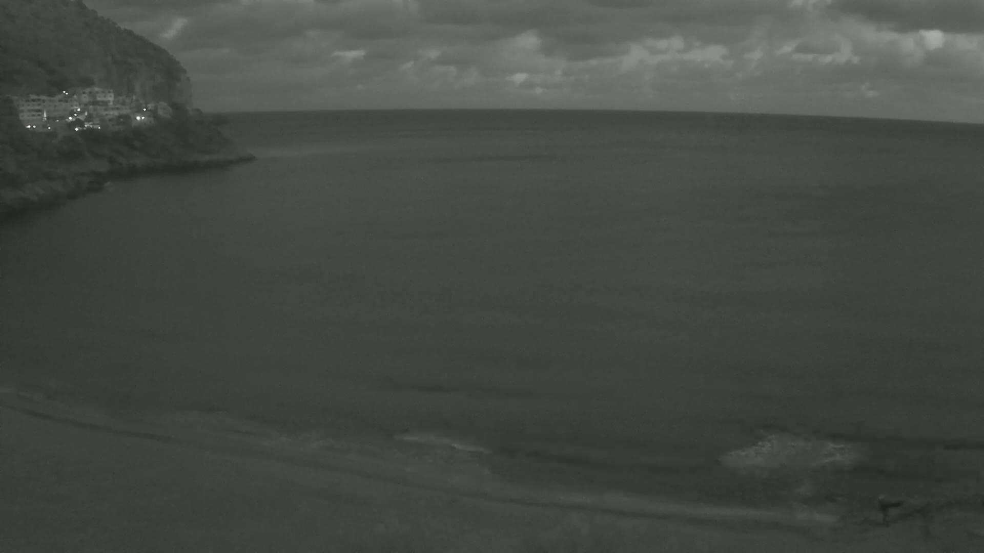 A dark, overcast sky hangs over a calm, gray sea lapping a sandy shore next to a rocky cliff with faint lights visible on its upper edge.