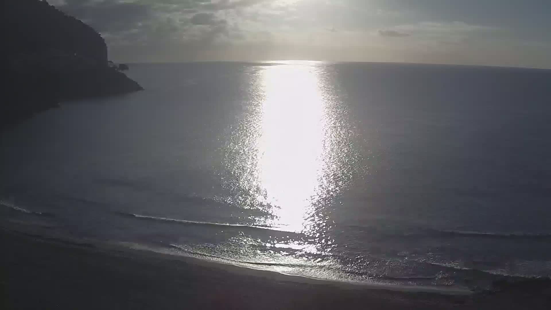 A bright sun shines on a calm ocean, reflecting on the water near a sandy shore and a dark cliff.